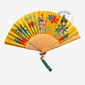 Once Upon Magic yellow Vaiana fan featuring Hei Hei and Pua with tropical flowers, wooden frame, and green tassel.