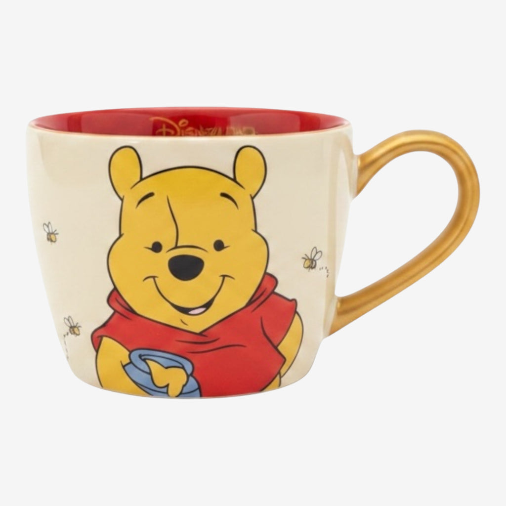 Tasse en céramique beige avec Winnie l'Ourson tenant un pot de miel, bordure rouge et anse dorée. Par Once Upon Magic.